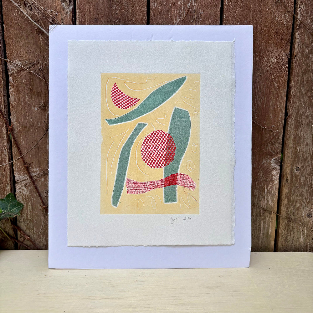 Framed abstract artwork with red and green shapes on a beige background, leaning against a wooden wall.