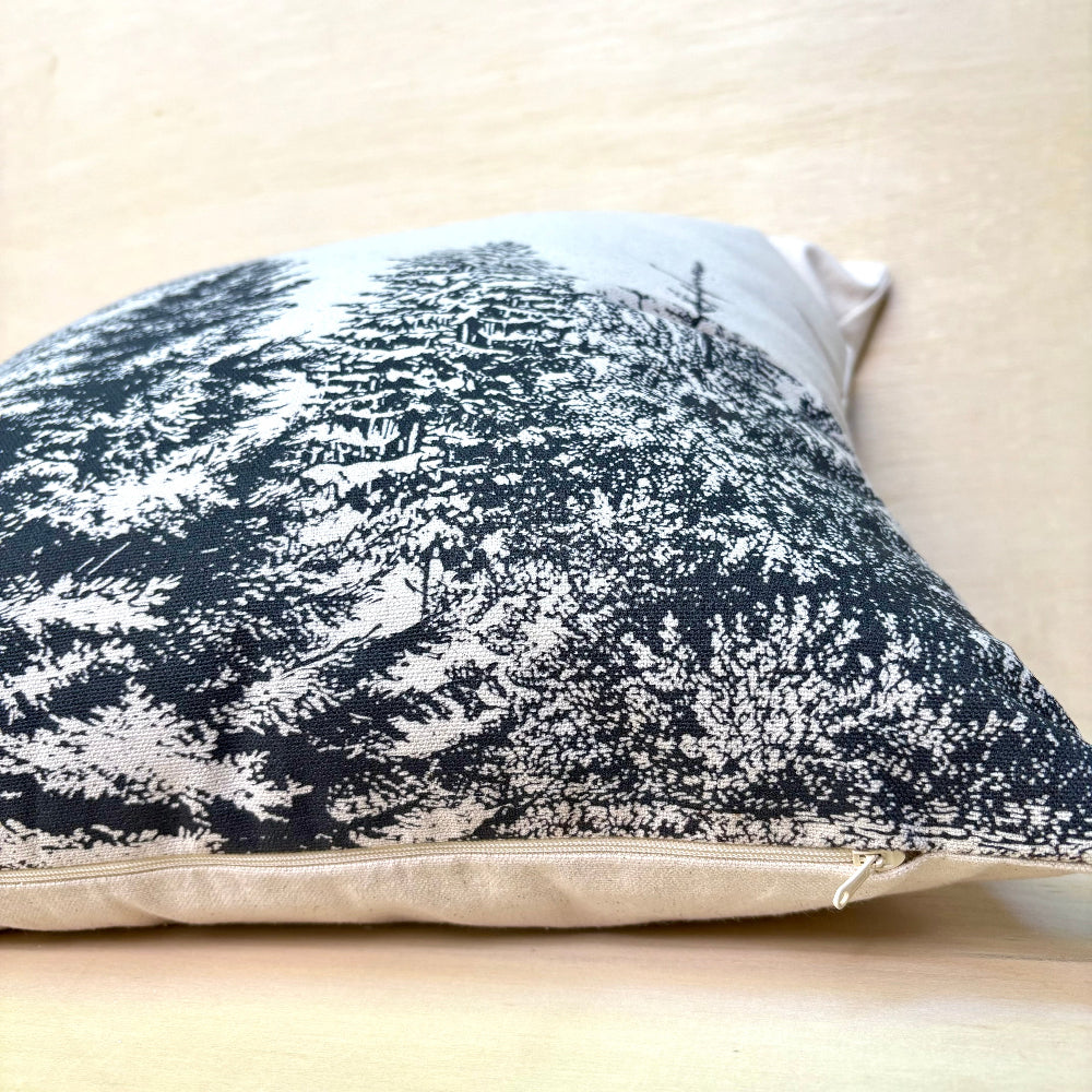 A square throw pillow with a black and white forest pattern, resting on a light beige wooden surface.
