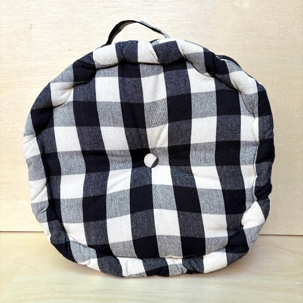 A round, tufted cushion with a black and white buffalo plaid pattern, featuring a top handle and a central button, set against a light wood background.