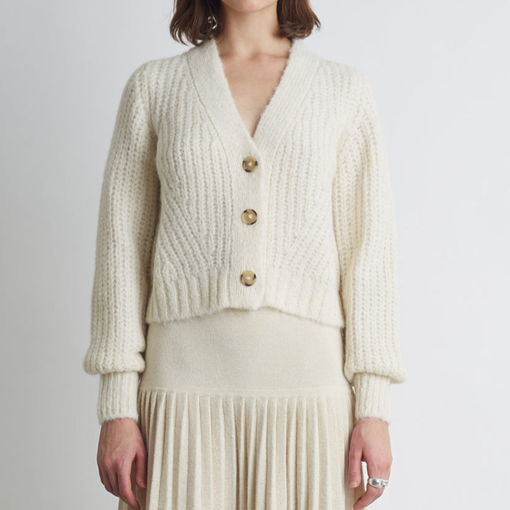 Woman wearing a cream-colored knitted cardigan with gold buttons on a plain background