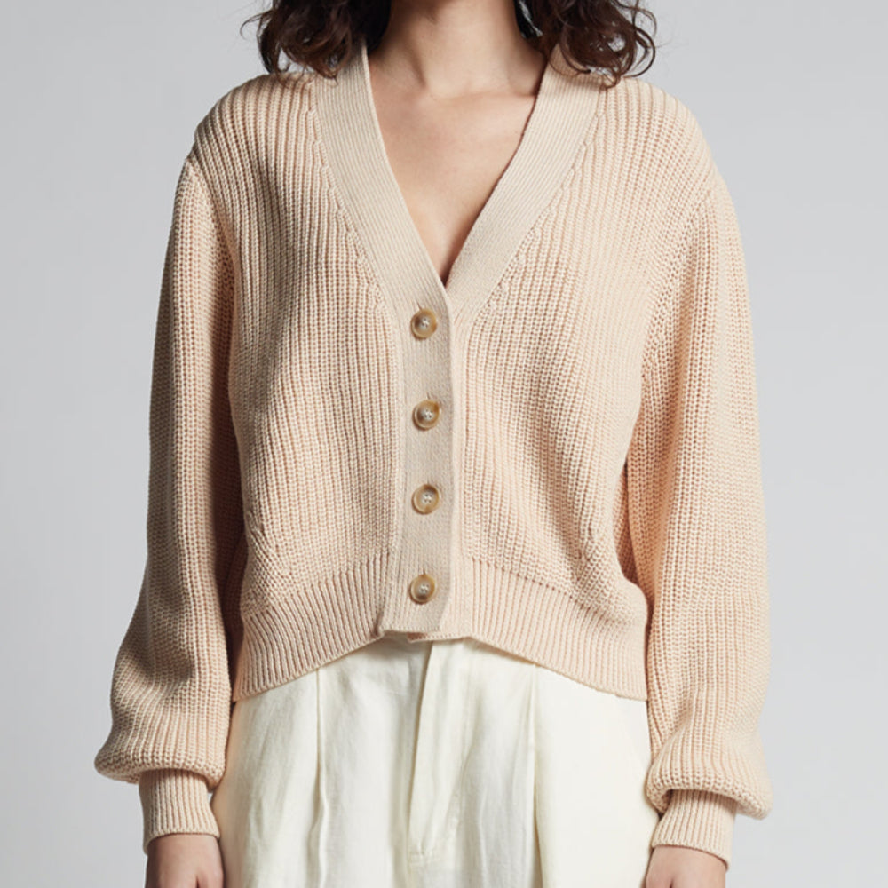 Woman wearing a beige cardigan over a beige top with white pants on a plain background