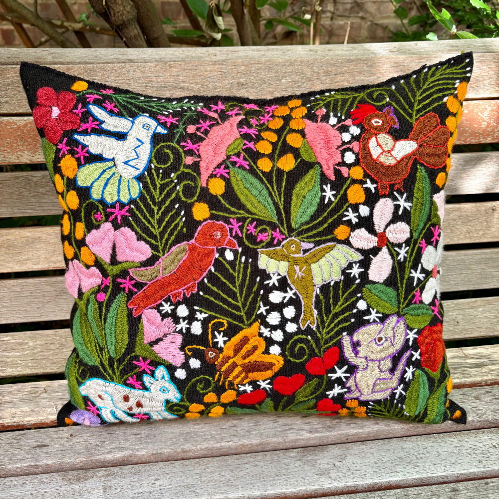 A square decorative throw pillow with a vibrant, hand-embroidered folk art design featuring birds, flowers, and butterflies on a black background, resting on a weathered wooden bench.