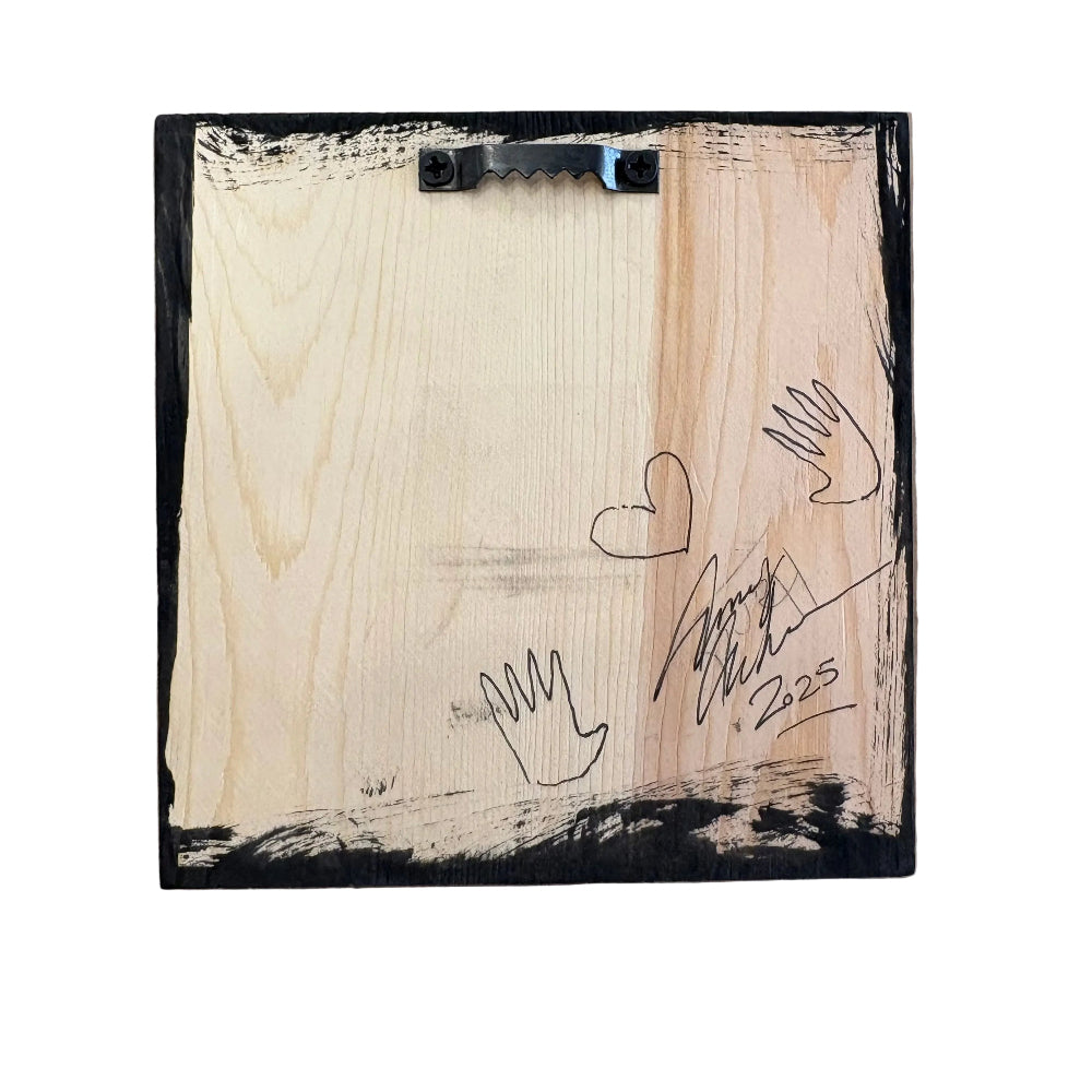 The back of a square wooden keepsake box or plaque, featuring a black metal hanger at the top, hand-drawn black ink sketches of hearts and hands, and a signature dated 2.205, set against a brown kraft paper background.