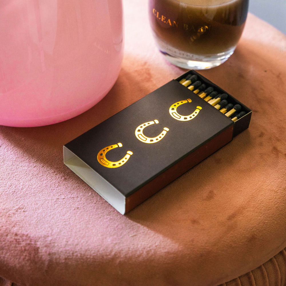 match box with gold horseshoes next to a candel and pink fase on a pink surface.