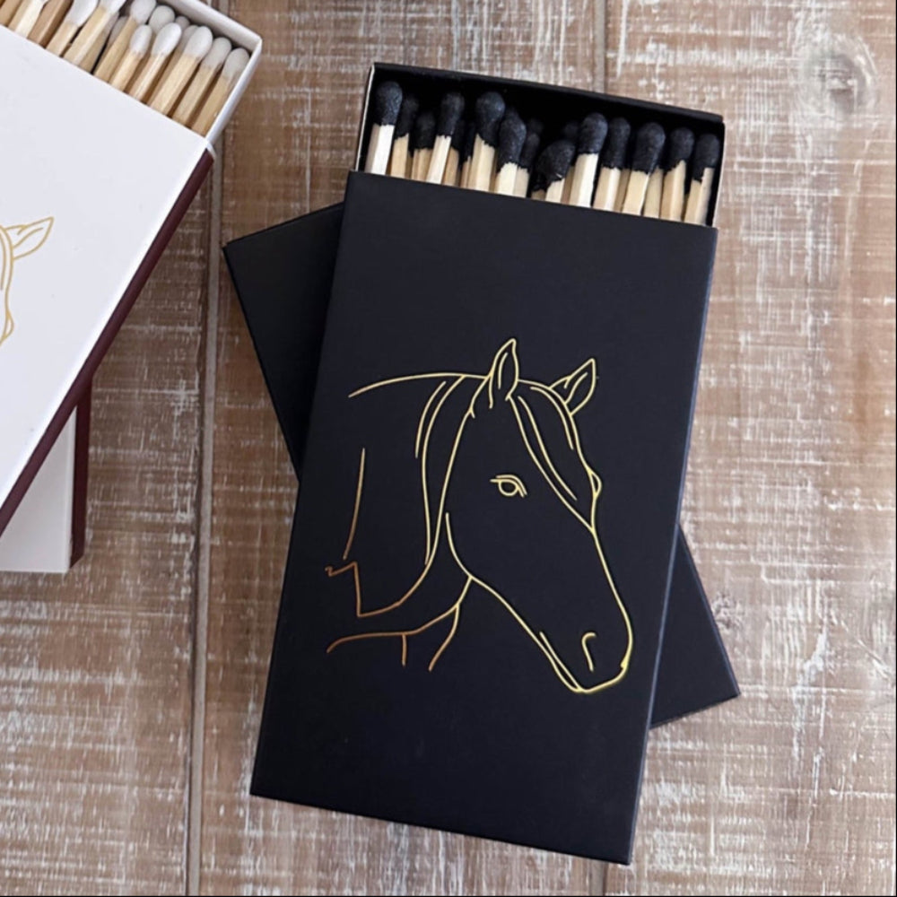Two matchboxes with horse illustrations on a wooden surface next to a candle.