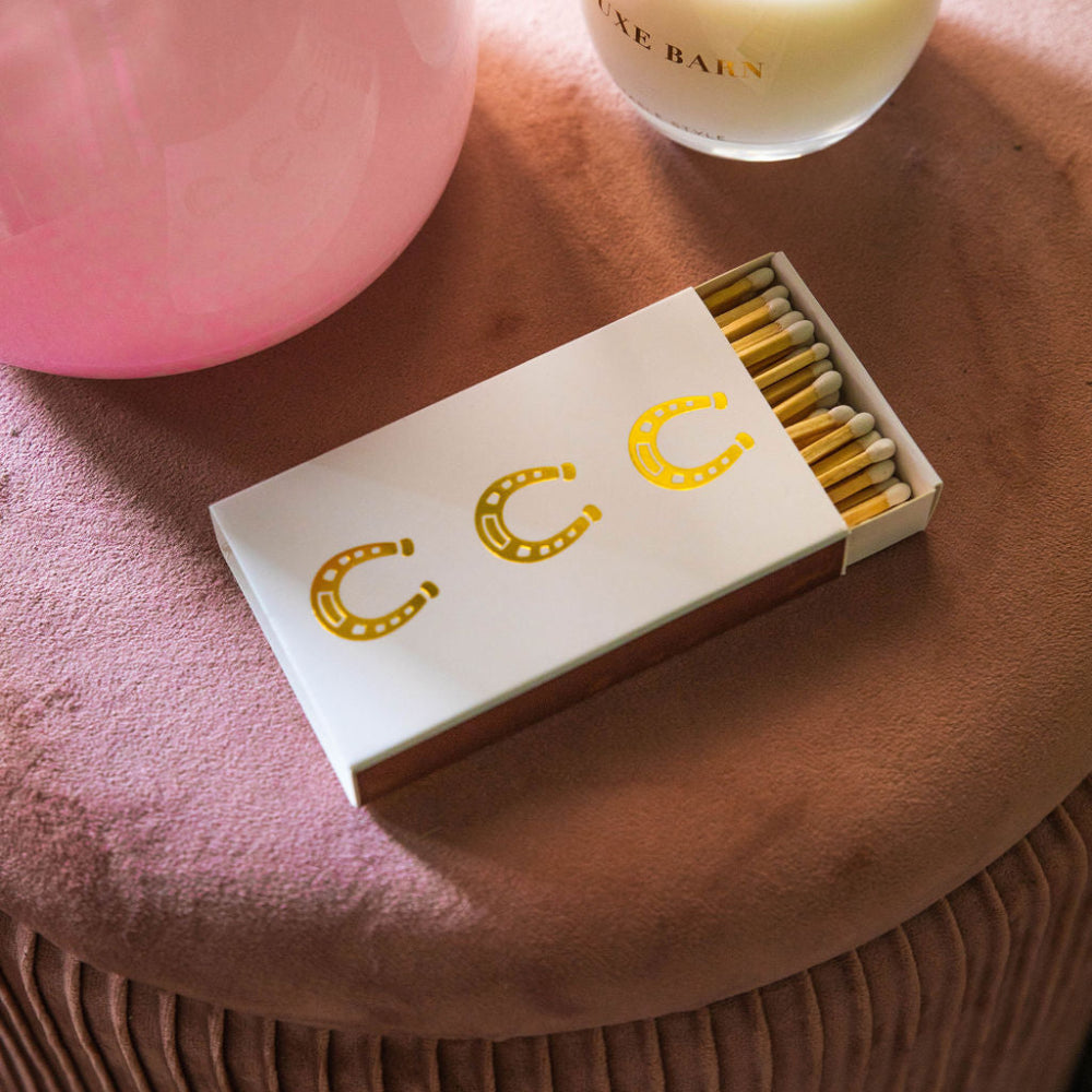 Matchbox with gold horseshoes next to a lit candle on a pink surface