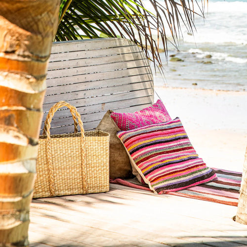 Beachside setting with a wooden bench, colorful pillows, and a woven bag under a palm tree.