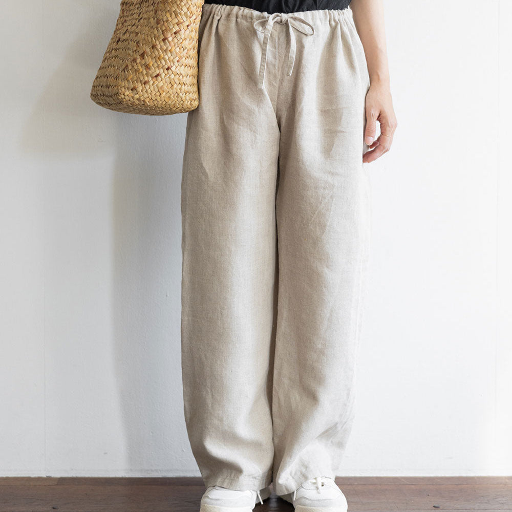 Person wearing a black top and beige pants holding a straw bag on a plain background