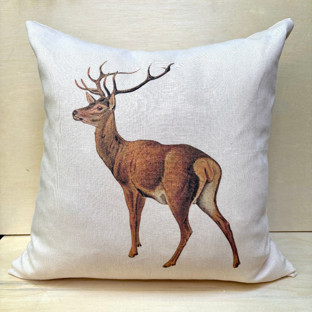 A square off-white linen throw pillow featuring a detailed illustration of a brown deer with antlers, set against a light wood background.