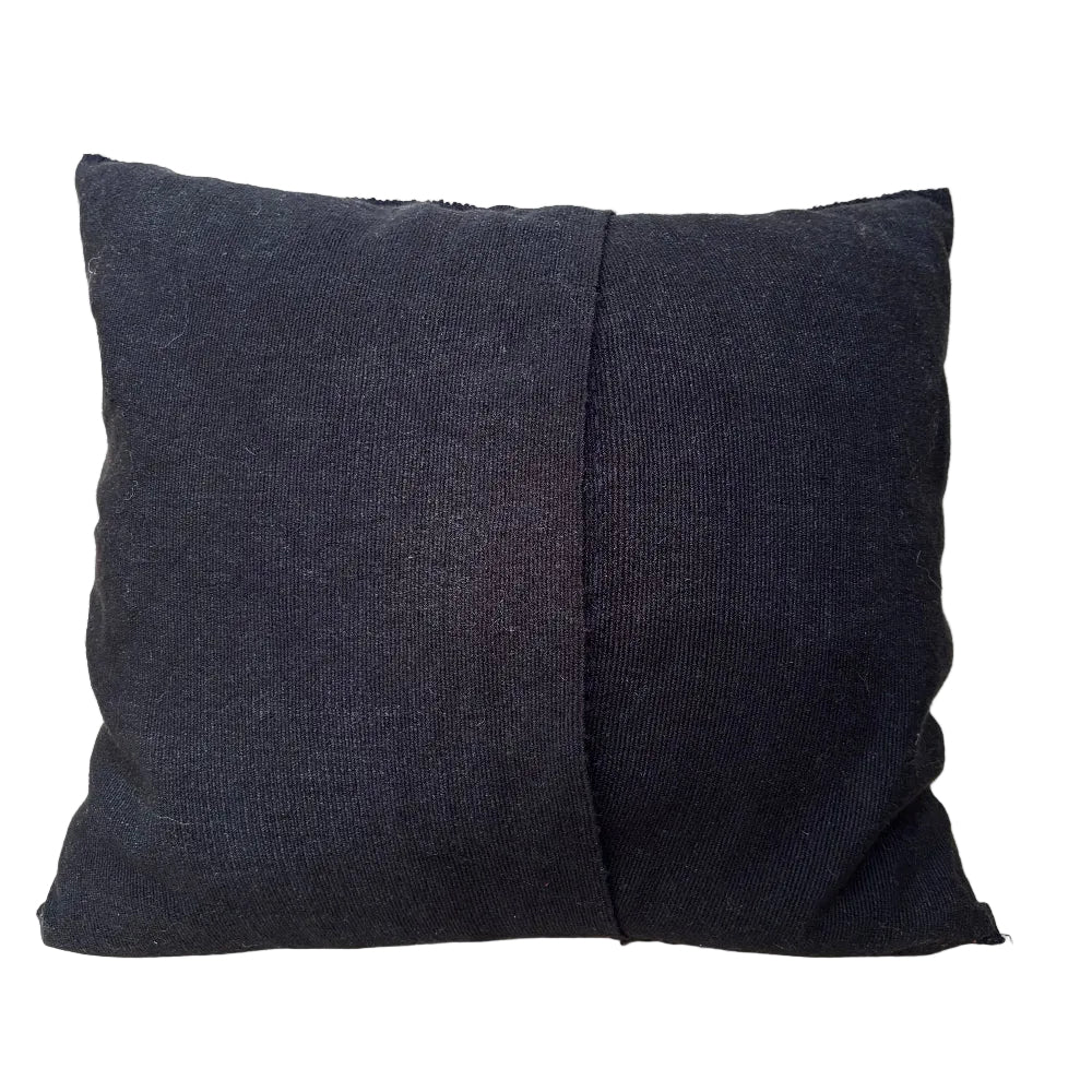 A square black textured throw pillow with a central seam, resting on a light-colored wooden surface against a light wood background.