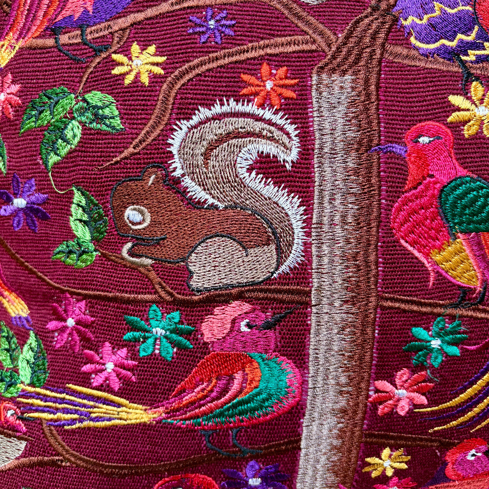 Close-up of an embroidered textile featuring a brown squirrel on branches with colorful birds, flowers, and leaves on a deep burgundy background.