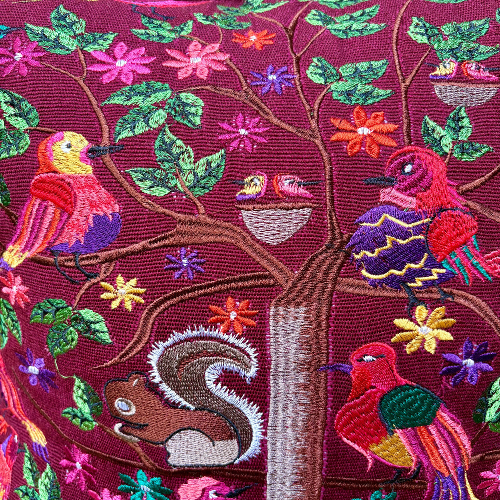 Close-up of an embroidered textile featuring a tree with colorful birds, flowers, leaves, and a squirrel on a deep burgundy background.