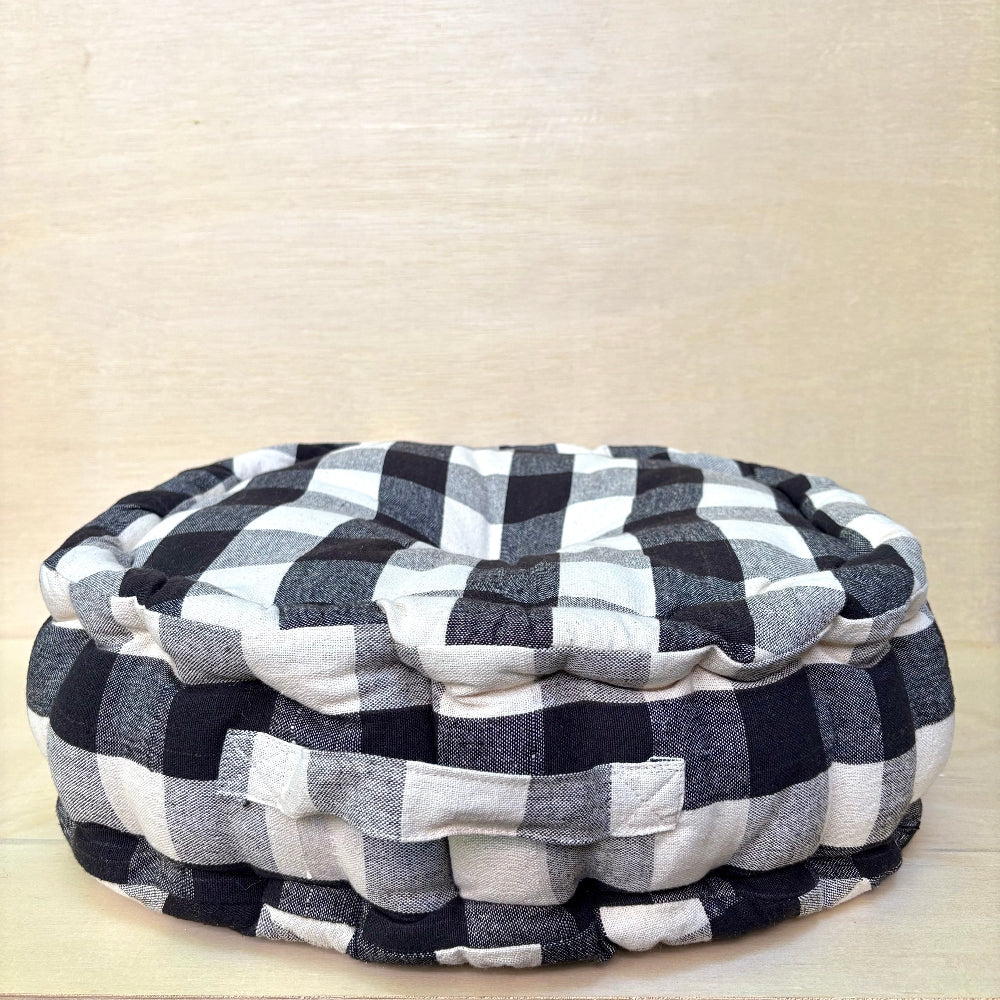 A round floor cushion or ottoman with a black and white buffalo plaid pattern, photographed against a light beige wood-grain background.