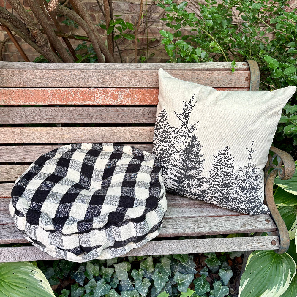 Two decorative cushions on a weathered wooden garden bench: a round black-and-white buffalo plaid pouf cushion and a square off-white throw pillow with a black pine tree print, set outdoors among green plants.