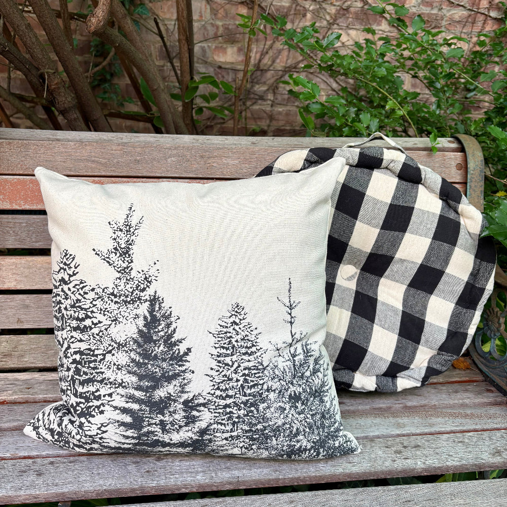 A square throw pillow with a black pine tree print on an off-white background, resting on a weathered wooden bench next to a black and white buffalo plaid blanket. A brick wall and green foliage are visible in the background.