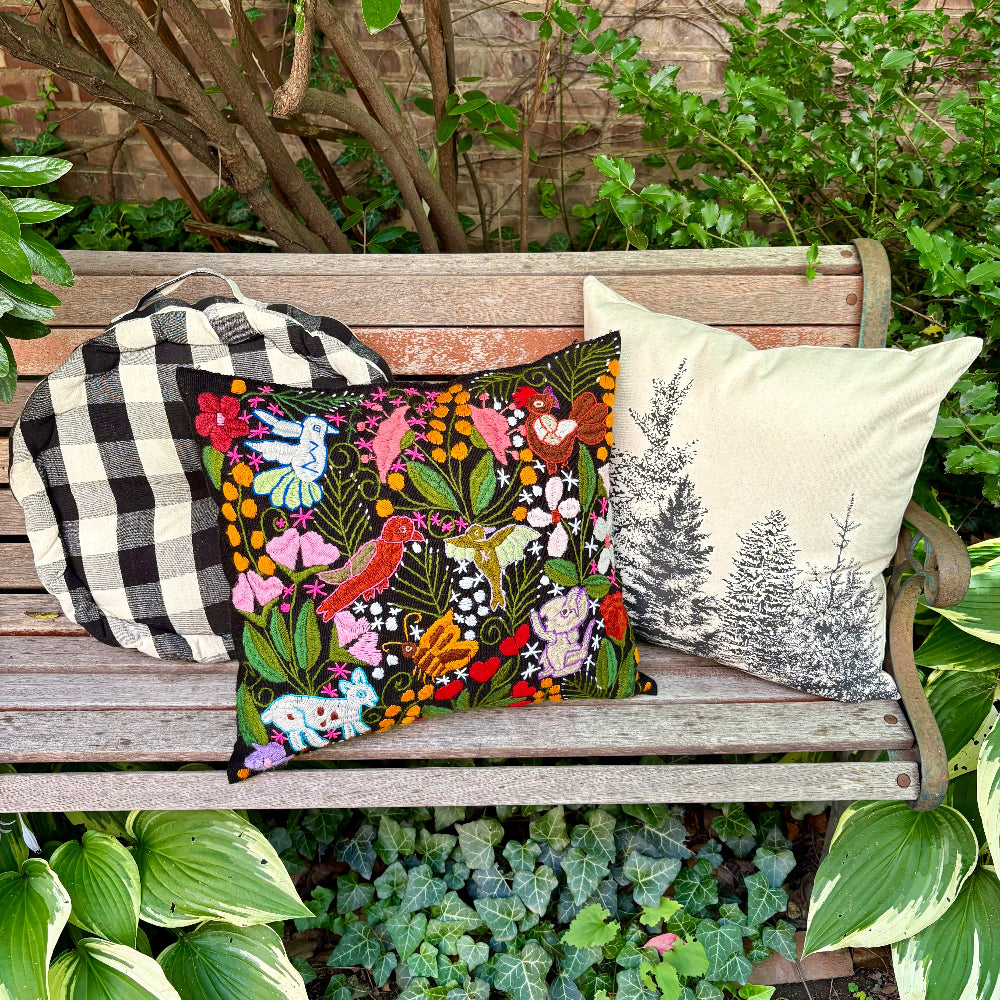 Three decorative throw pillows arranged on a wooden garden bench surrounded by green foliage. The center pillow features a colorful embroidered floral and bird pattern, the left pillow is black and white gingham, and the right pillow is cream with a black pine tree print.