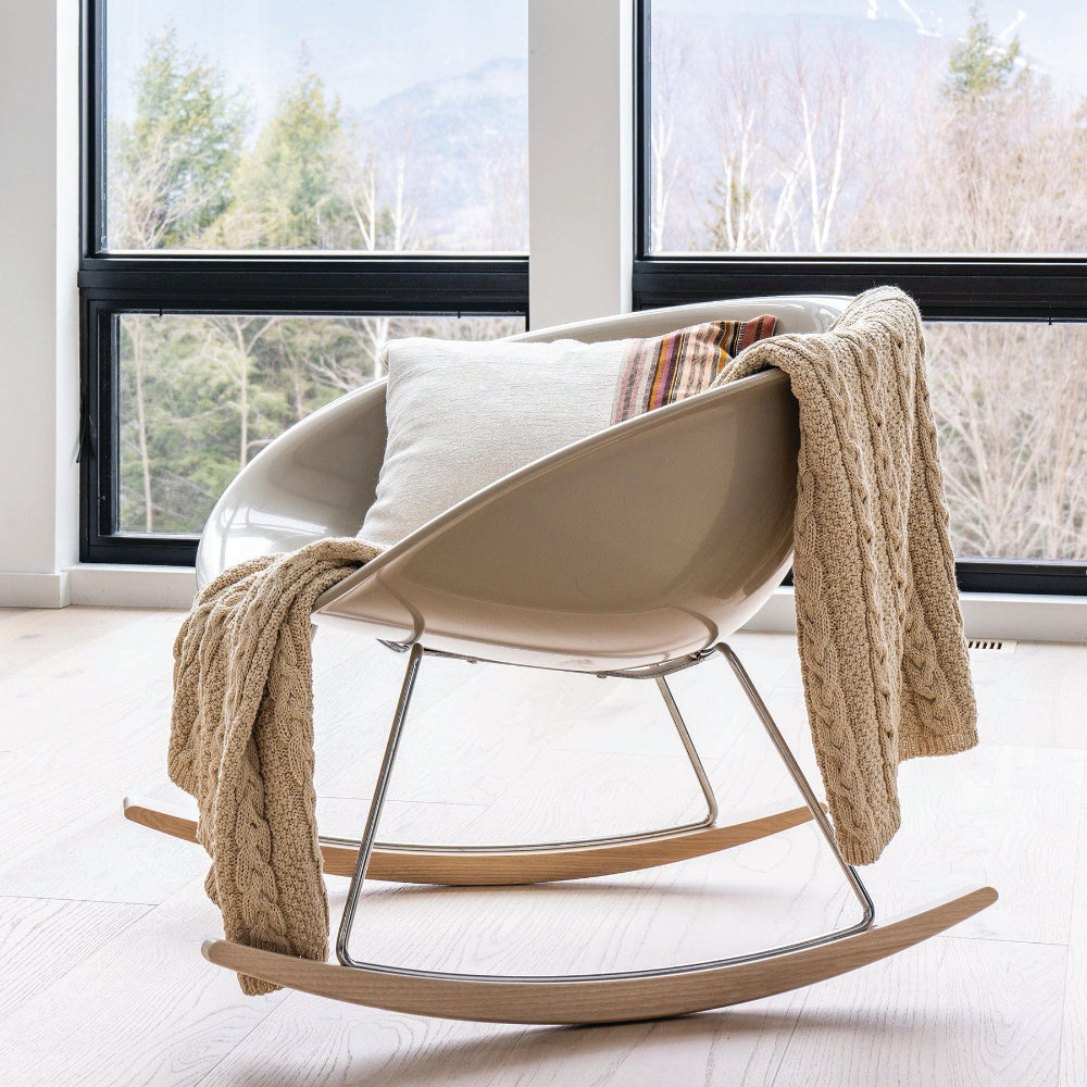 Beige modern rocking chair with a blanket and pillow in a room with large windows.