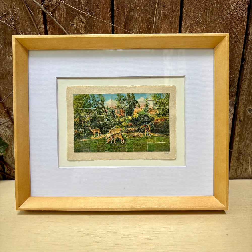 Framed artwork of deer in a natural setting on a wooden surface.