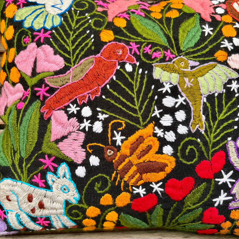 Close-up of a black embroidered fabric featuring colorful floral and insect motifs, including pink flowers, green leaves, a red bird, a yellow-green hummingbird, an orange-and-brown butterfly, and a white rabbit with blue outlines.