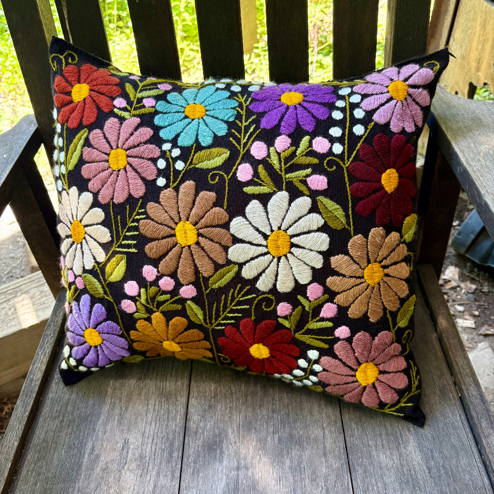 A square decorative throw pillow with colorful embroidered flowers on a black background, resting on a weathered wooden garden chair.