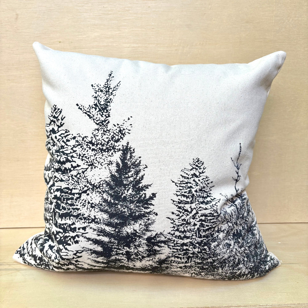 A square throw pillow with a black pine tree forest pattern on a light beige linen background, resting on a light wood surface.
