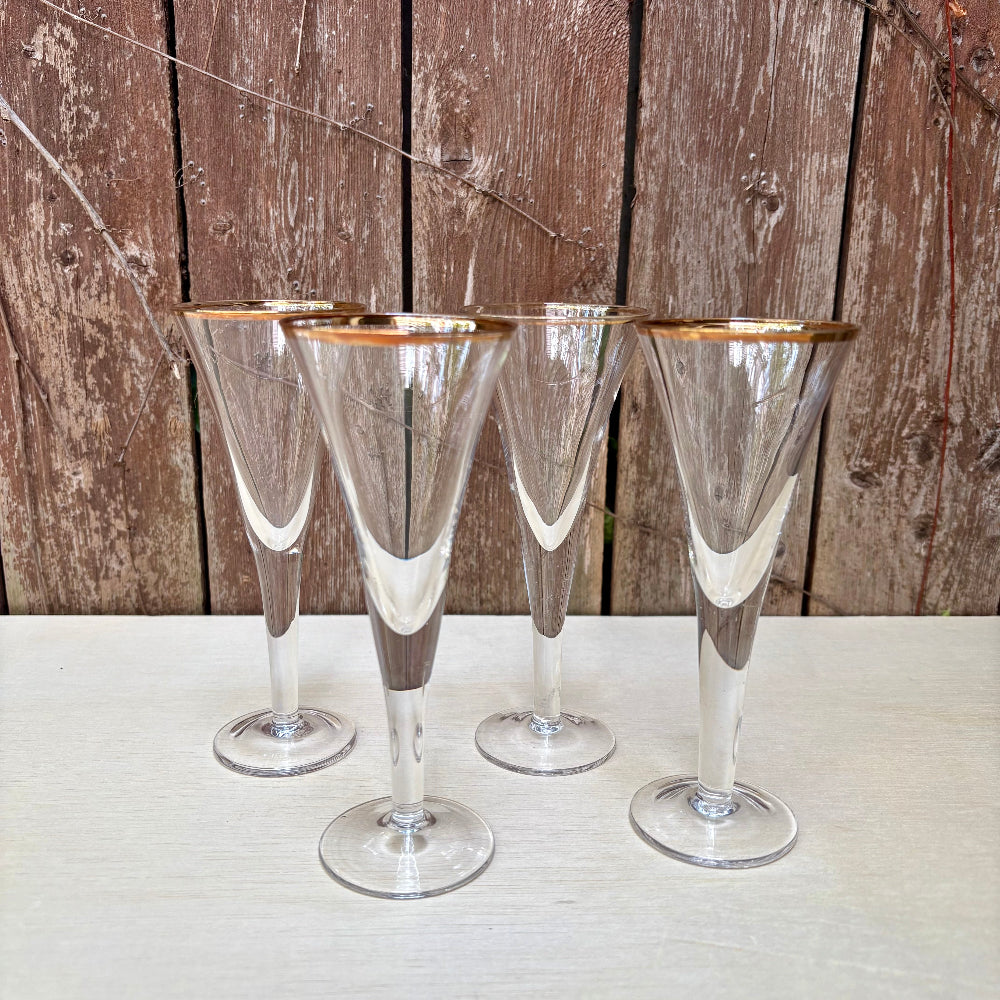Four cordial flutes with gold rims arranged on a light tabletop against a rustic brown wooden plank background.