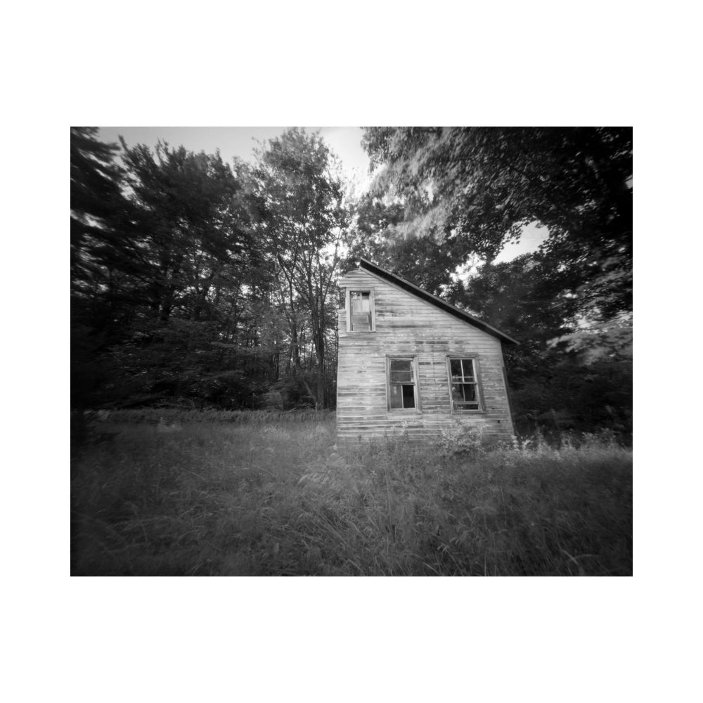Black and white photograph of an old weathered wooden cabin surrounded by tall grass and dense trees, presented as an archival art print with a white border.