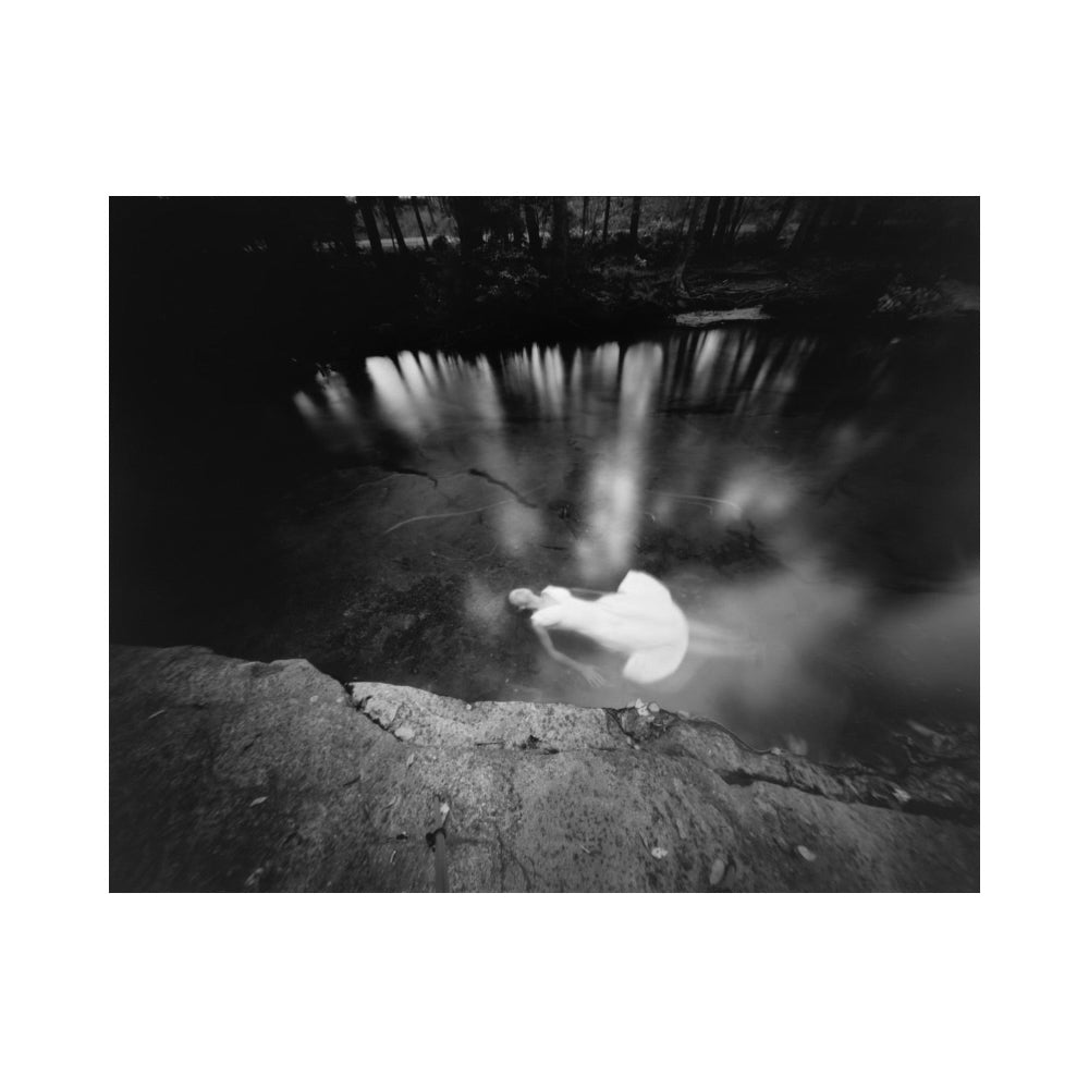 Black and white fine art photograph of a woman in a flowing white dress standing in a dark, reflective body of water, framed with a wide white border.