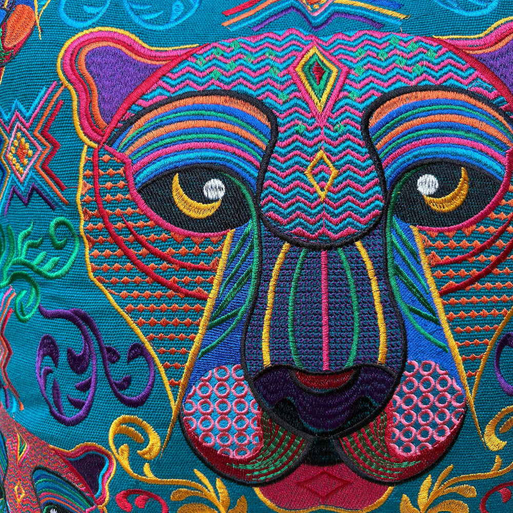 Close-up of a colorful embroidered animal face, likely a lion or similar big cat, with intricate geometric and floral patterns in turquoise, purple, pink, yellow, green, and orange on a patterned textile background.