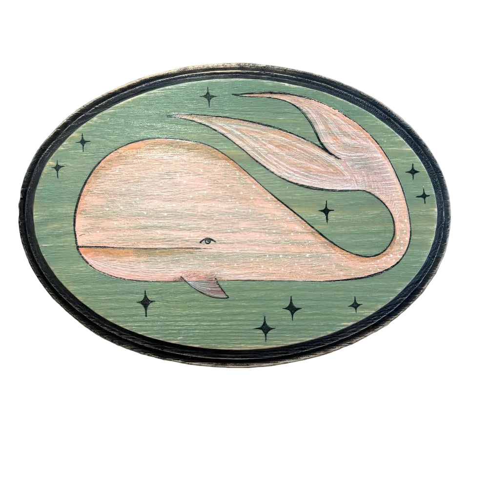 An oval wooden plaque featuring an inlaid whale design on a sage green background with black star accents, photographed on a brown textured surface.