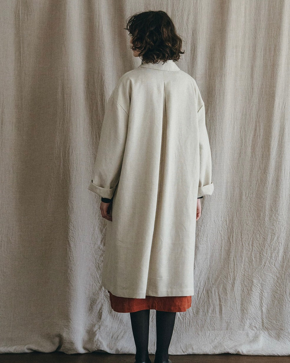 Person wearing a long beige coat standing against a textured white curtain.